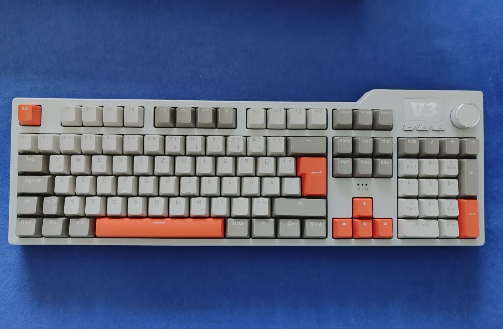 Hell-graue mechanische Tastatur AJAZZ AK35I V3 mit orangefarbenen Keycaps, Drehregler links und V3-Schrift auf dem oberen Bereich.